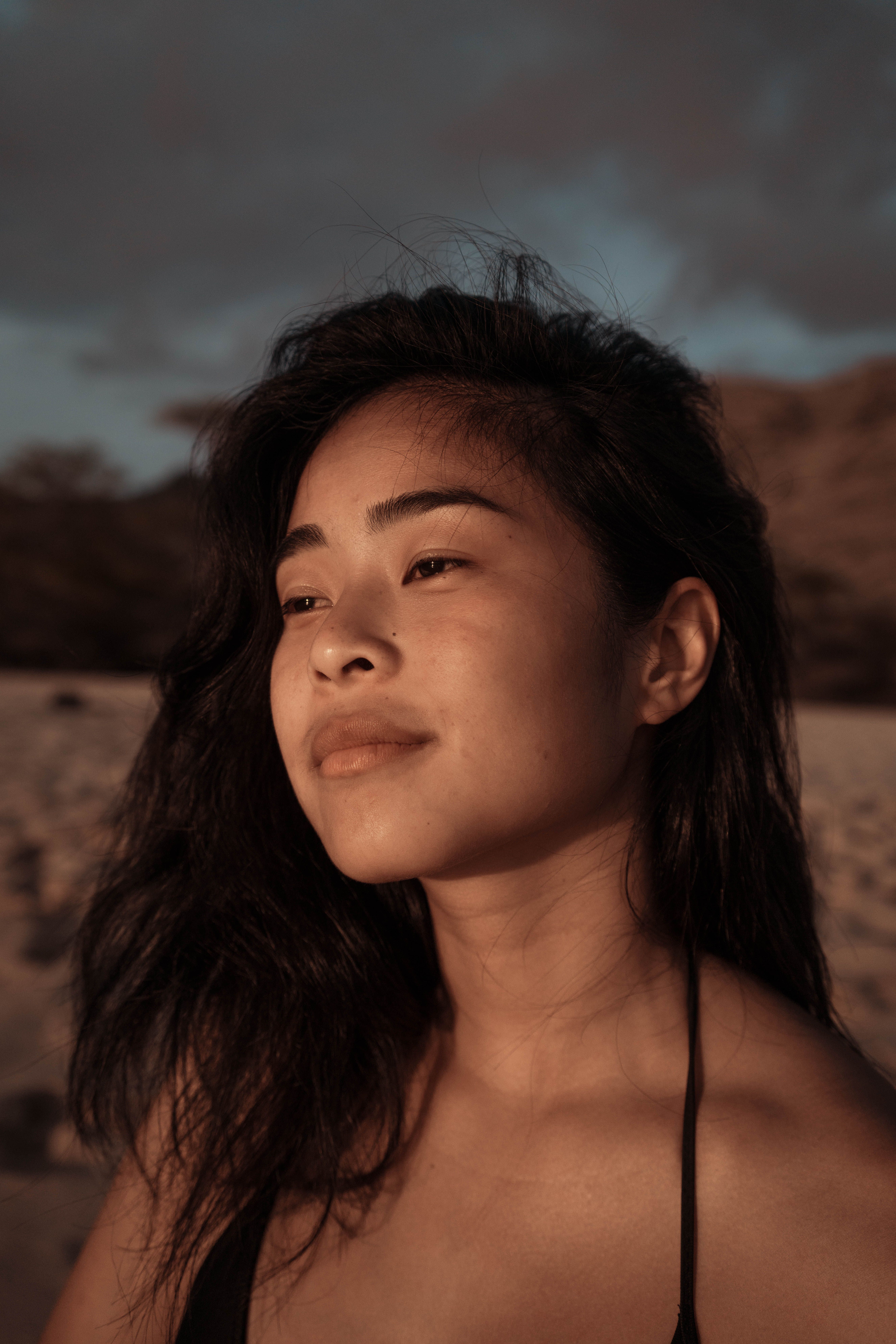 A contemplative young woman looks afar on a beach during sunset, capturing a serene moment.