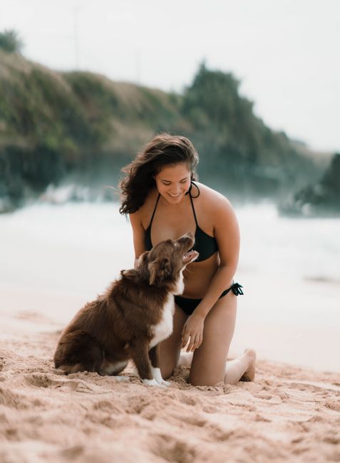 A woman in a bikini happily plays with her dog on a sandy beach.