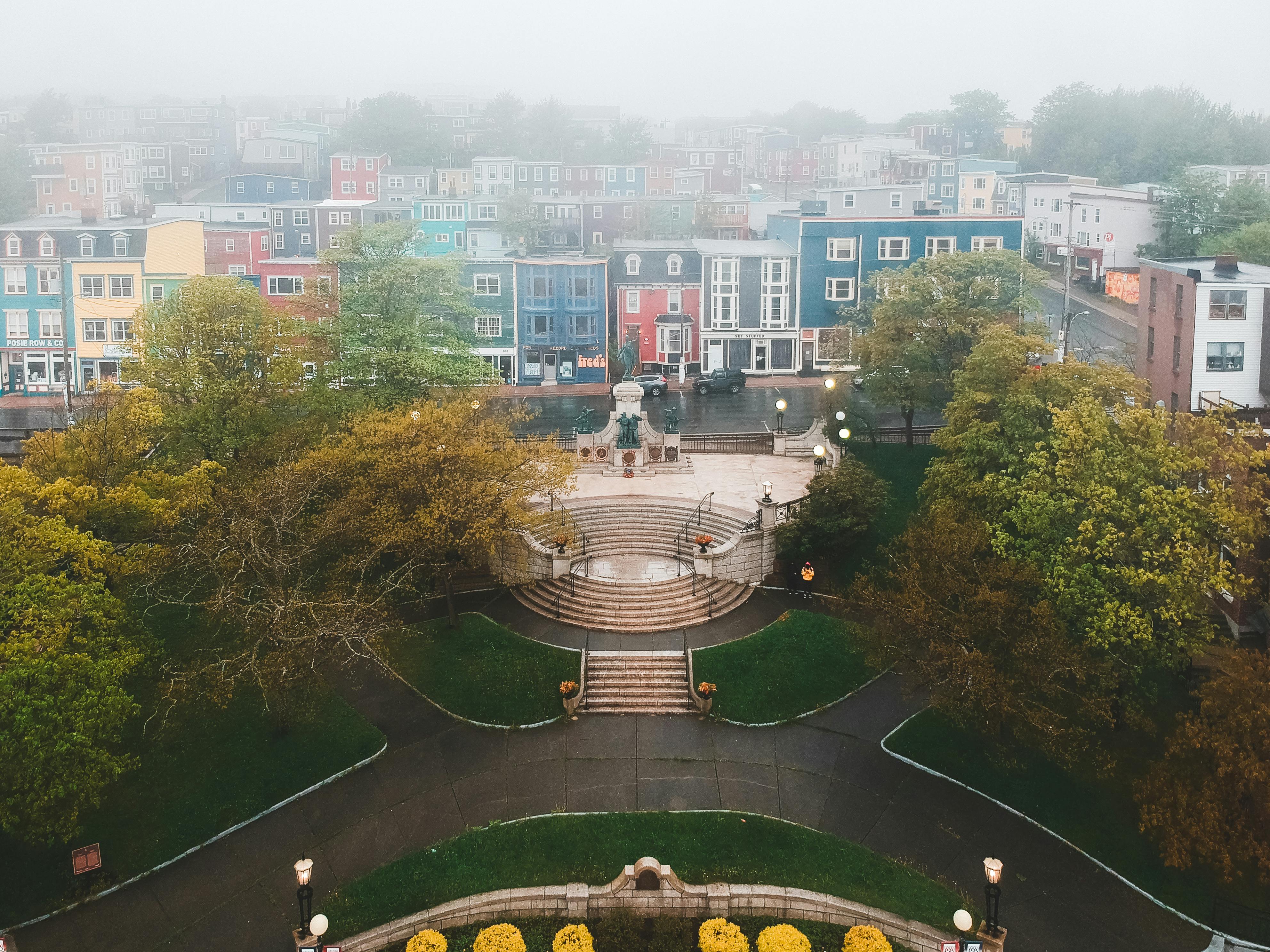 From above of aged stairs and statues near trees behind colorful residential building exteriors under sky in fog