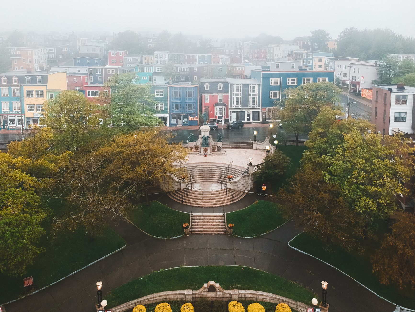 From above of aged stairs and statues near trees behind colorful residential building exteriors under sky in fog