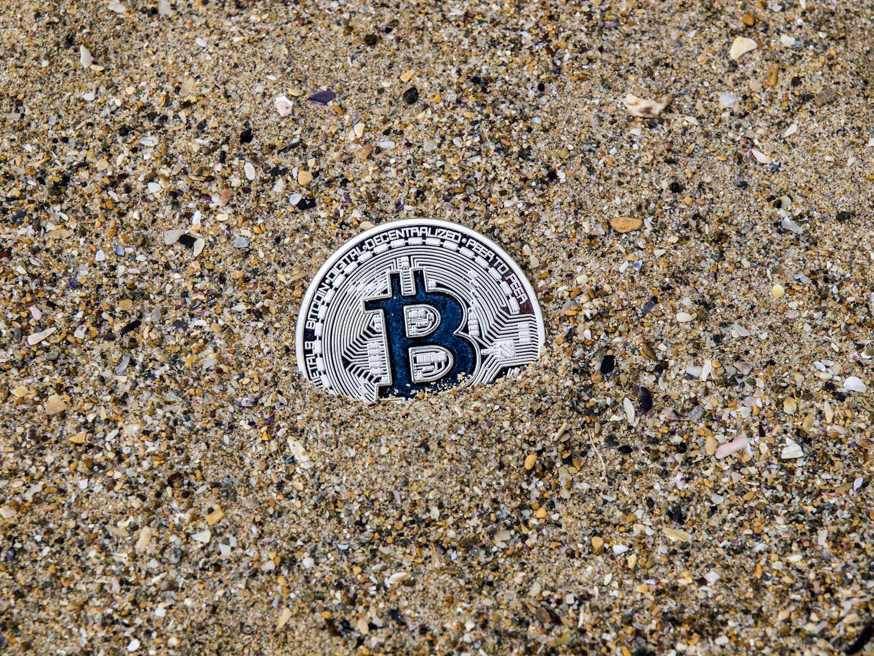 A conceptual image of a Bitcoin symbol partially buried in sand, symbolizing digital currency.
