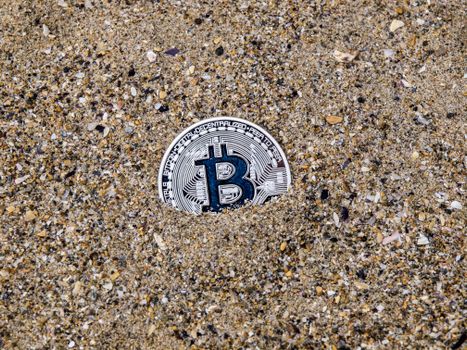 A conceptual image of a Bitcoin symbol partially buried in sand, symbolizing digital currency.