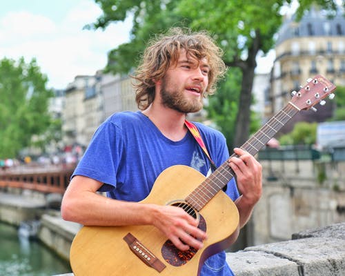 Positive young male musician in casual wear playing acoustic guitar and singing while standing on old city street on sunny summer day