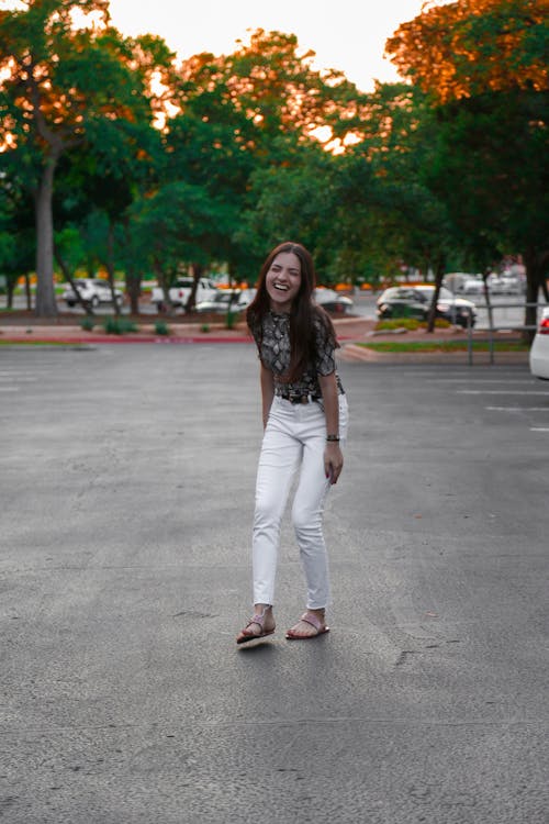 Full body young excited brunette in trendy white trousers and gray blouse standing in city park and laughing happily