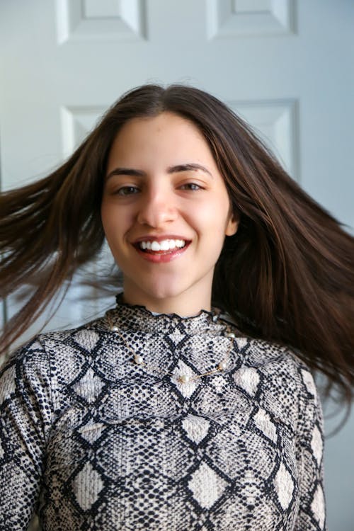 Cheerful casual young lady with long brown hair smiling and looking at camera against white wall