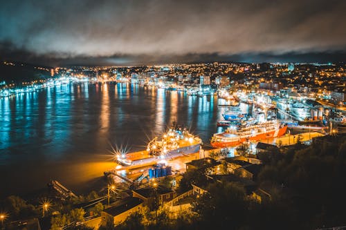 From above of illuminated city with typical buildings and port with moored ships located on seashore against cloudy night sky