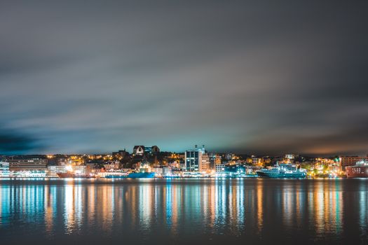 在多云的夜空下，圣约翰市的灯光倒映在漂浮于海面的船只上，构成一幅如画的风景