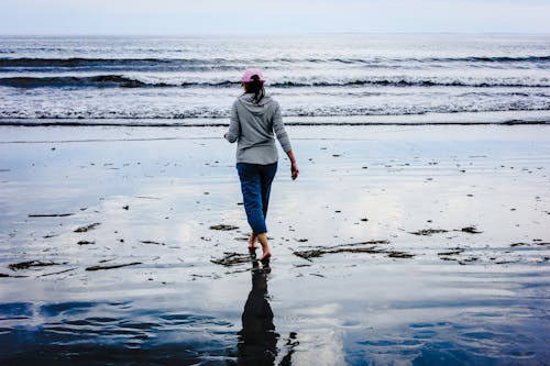 Back view of anonymous barefooted female traveler in casual clothes and cap walking on wet sandy beach near waving ocean