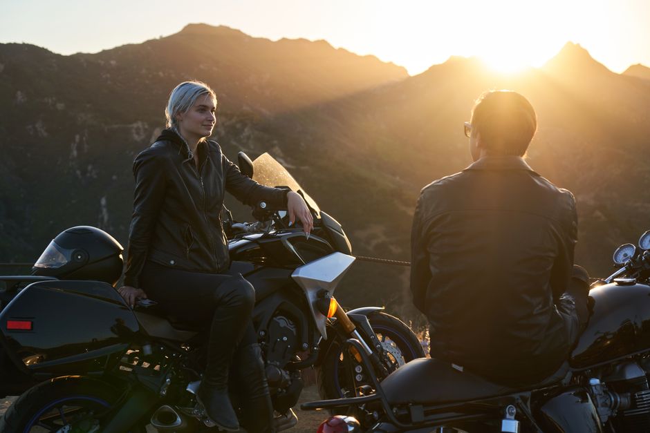 A biker couple wearing leather jackets, sitting on motorcycles at sunset with mountains in the background.