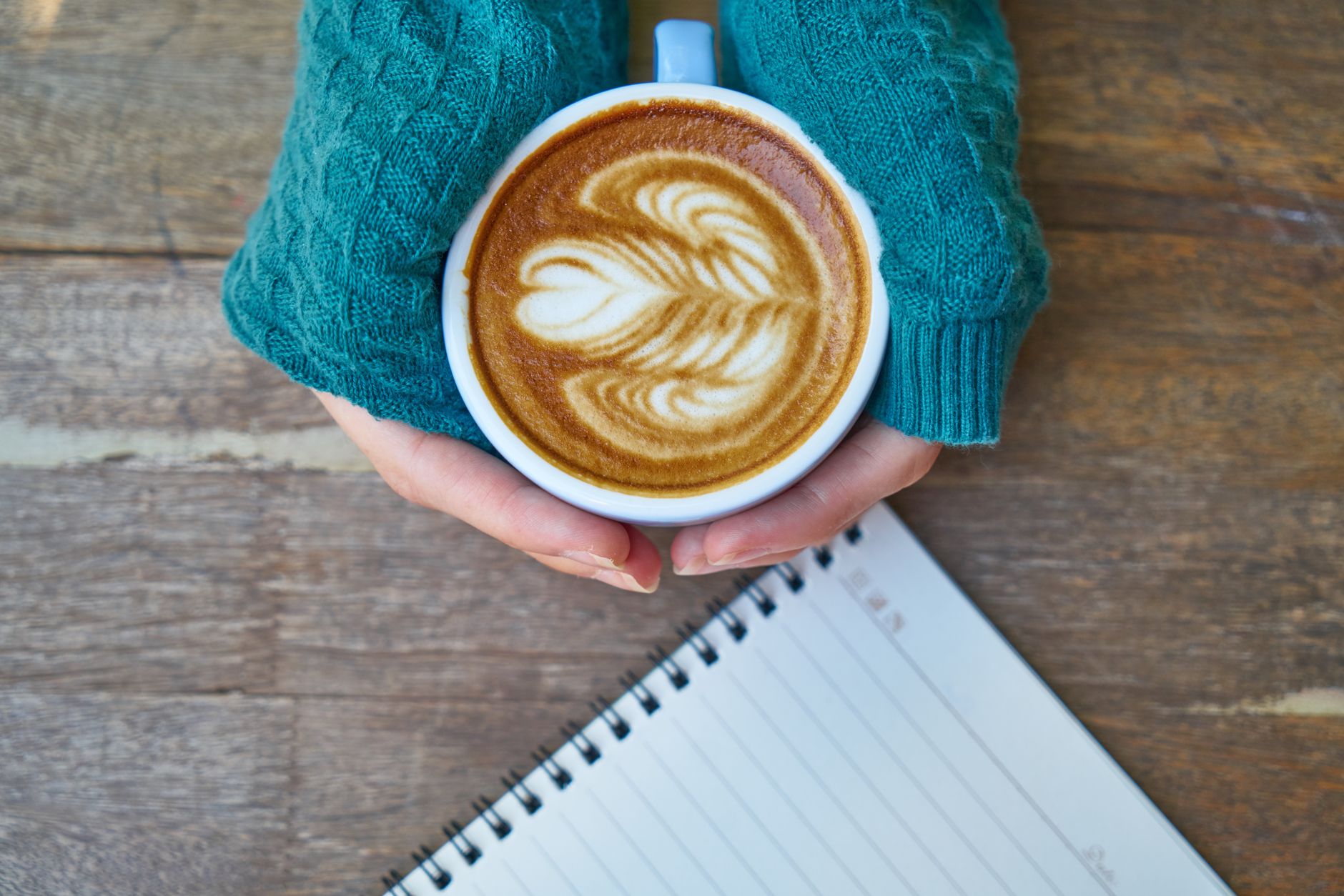 A person holds a cup of latte with beautiful art on a wooden table next to a notebook.