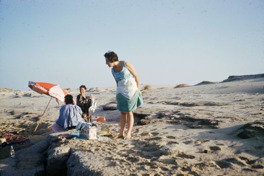 A classic vintage beach scene with women enjoying a picnic under a parasol.