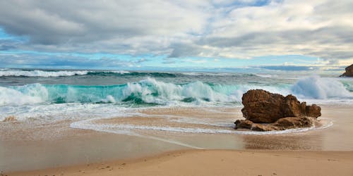 Magnificent scenery of stormy ocean waves washing sandy coast with rough rock against cloudy sky