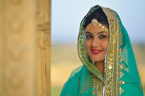 Gorgeous young ethnic lady with red lips in traditional sari and ghoonghat headscarf standing against blurred background and looking at camera