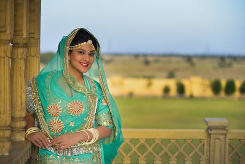 Content young Indian lady in green ornamental sari and ghoonghat resting on terrace of oriental palace on sunny day