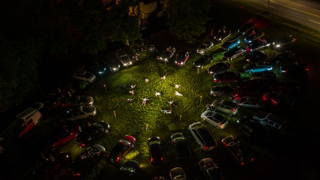 Drone view of anonymous people lying on green lawn surrounded by various cars with glowing headlights at night