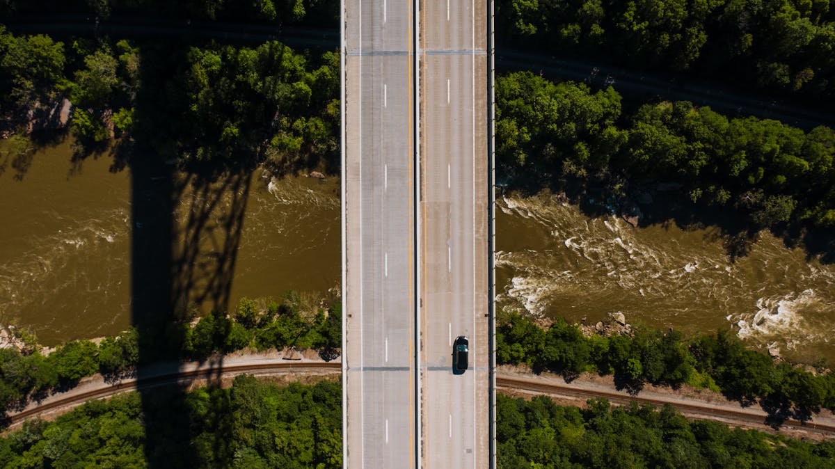 Aerial view of lonely car driving along asphalt bridge road crossing wild river running through green forest
