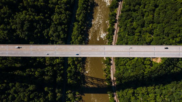 Breathtaking drone view of modern bridge road with driving cars crossing brown river flowing between lush green woods on sunny day