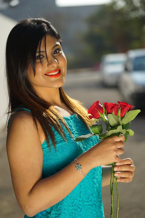 Side view of ethnic female in dress standing with bunch of red roses and smiling at camera