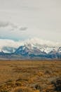 Brown Grass Field Near Snow Covered Mountains Under White Cloudy Sky