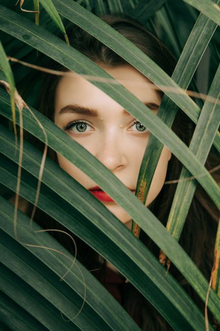 Close-up portrait of a woman with red lips peeking through lush green palm leaves.