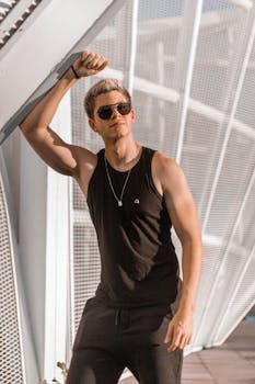 Serious male in black outfit with sunglasses looking away while standing near metallic construction with raised hand during daytime on street