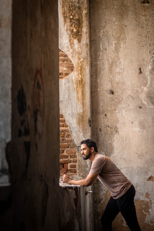 Side view of concentrated male leaning on windowsill and looking away while standing in abandoned building with shabby walls during daytime