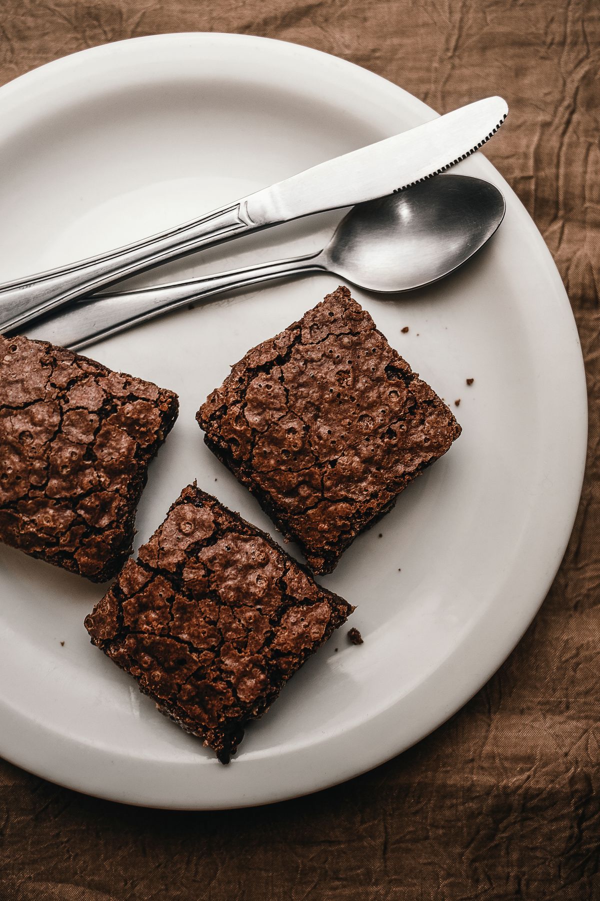 Three square brownies with shiny crackled tops on a white plate with knife and spoon