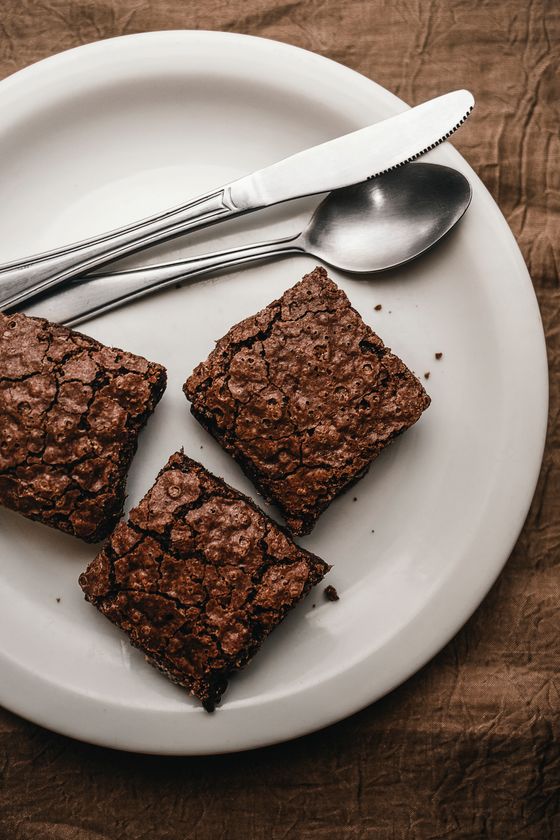Three square brownies with shiny crackled tops on a white plate with knife and spoon