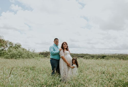 Free Content chubby male embracing expectant girlfriend in dress near charming girl on grass lawn in countryside while looking at camera Stock Photo