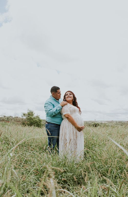 Free Side view of cheerful chubby male embracing stylish pregnant girlfriend in dress on grass lawn under cloudy sky while looking at each other Stock Photo