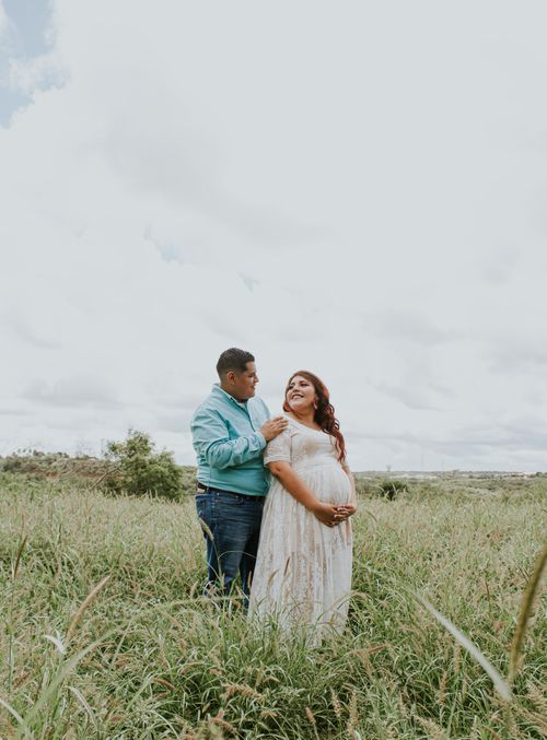 Free Smiling male embracing elegant expectant partner on grass meadow under cloudy sky while looking at each other Stock Photo