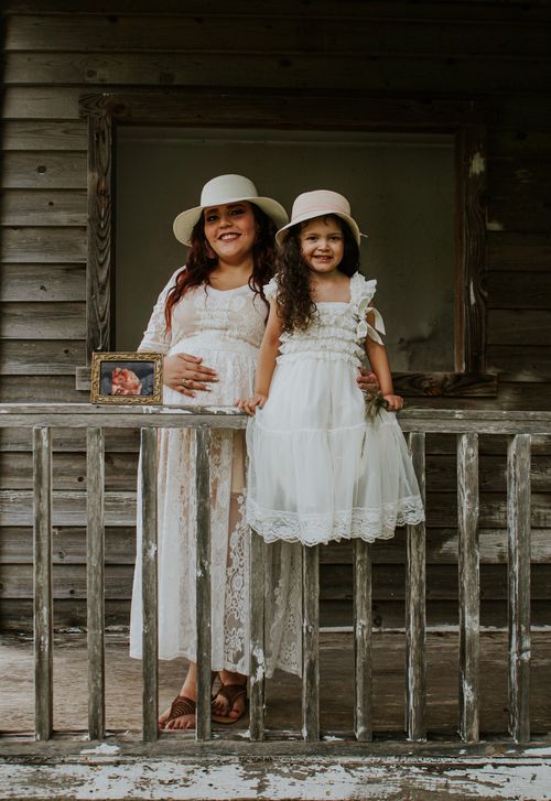 Free Cheerful expectant woman in straw hat embracing smiling girl on porch near fence with photo of fetus while looking at camera Stock Photo