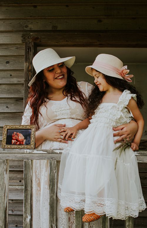 Free Cheerful woman in straw hat embracing content daughter in white dress touching belly on fence of terrace near photo of fetus Stock Photo