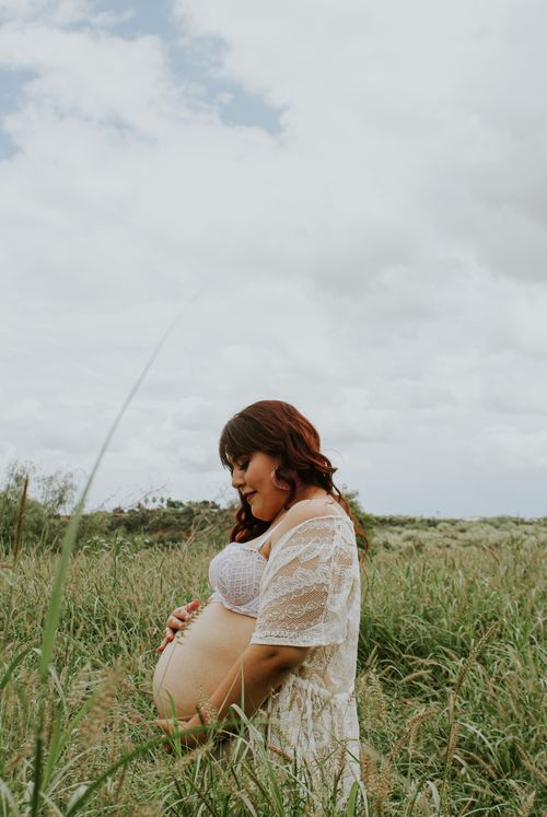 Free Side view of smiling expectant female in brassiere touching belly on grass meadow under cloudy sky Stock Photo