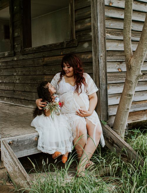 Free Cheerful expectant mother in dress embracing charming little kid while sitting on wooden porch and looking at each other Stock Photo