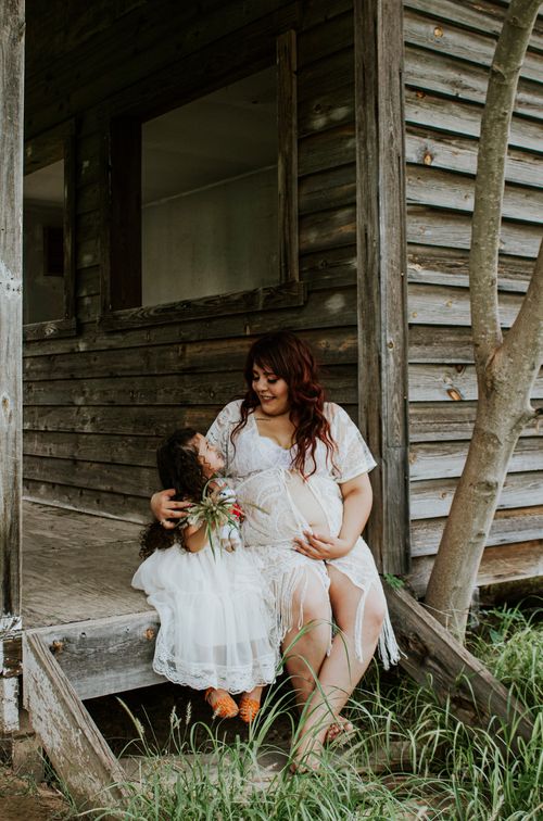 Free Cheerful pregnant female touching tummy while embracing charming daughter sitting together on wooden terrace and looking at each other in countryside Stock Photo