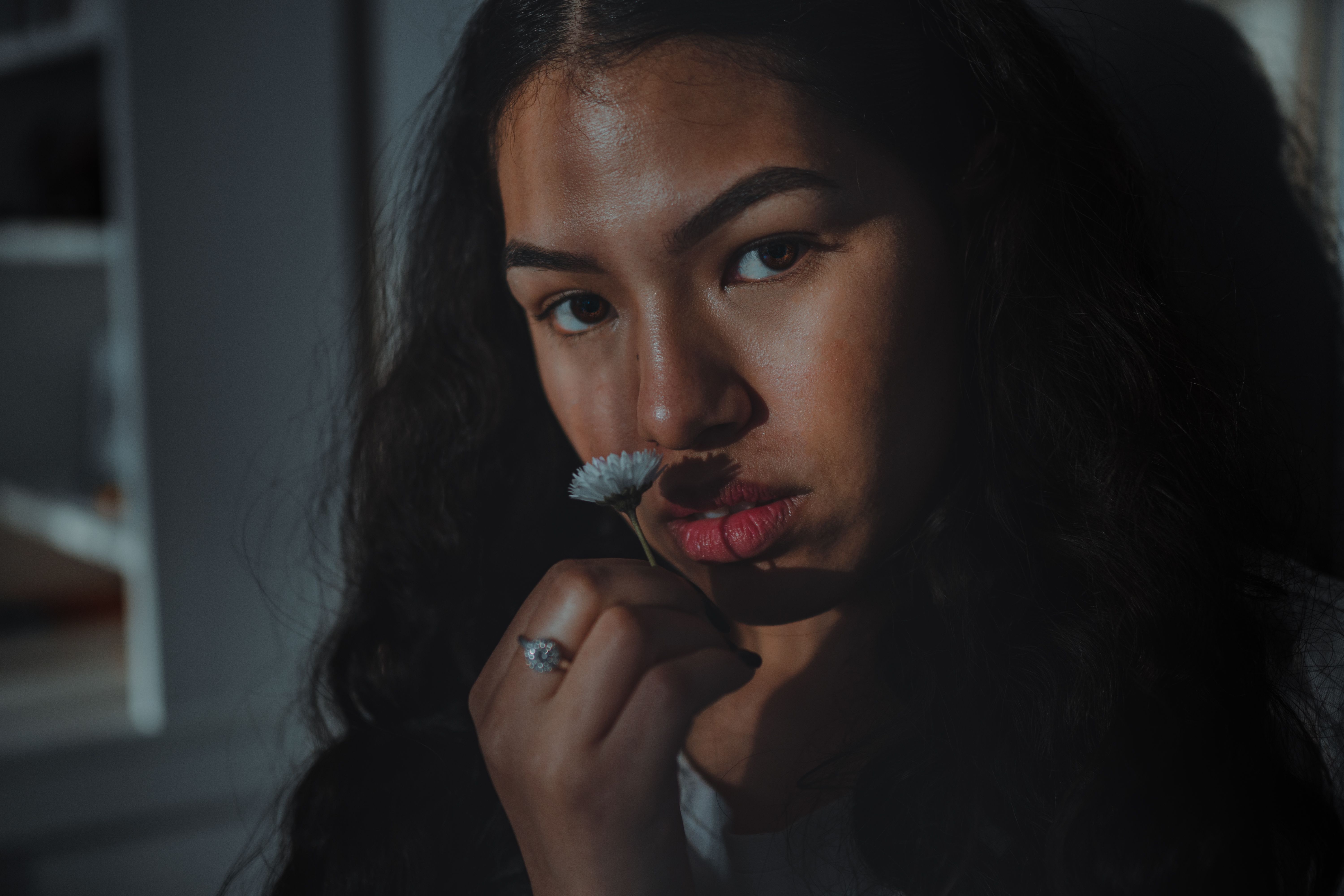 Close-up portrait of a woman with a moody expression holding a flower indoors.