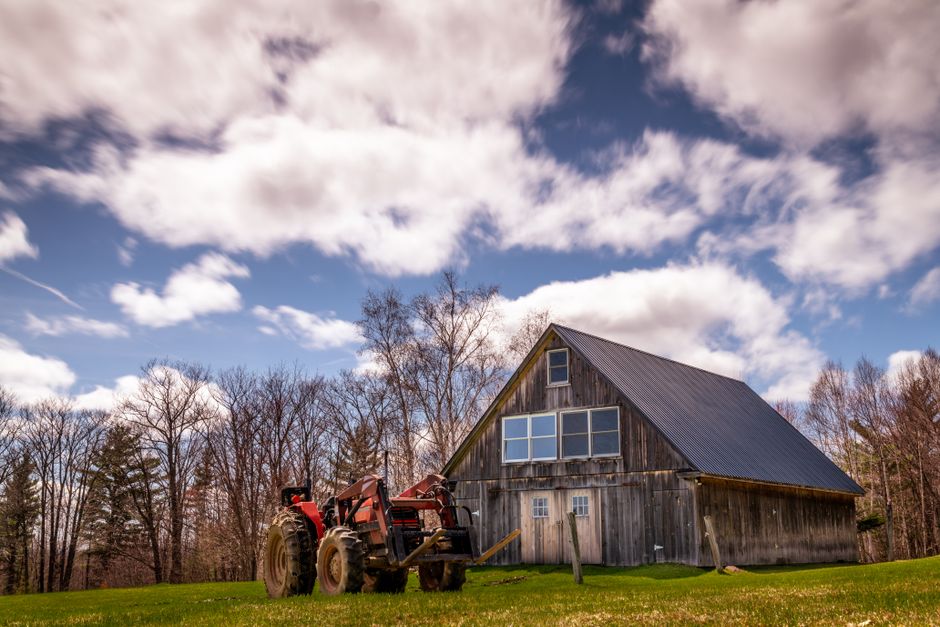 Scenery of abandoned barn located near old tractor on grassy lawn amidst leafless trees in countryside