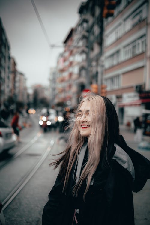 Delighted Asian female in black warm hoodie standing on busy city street with sincere smile