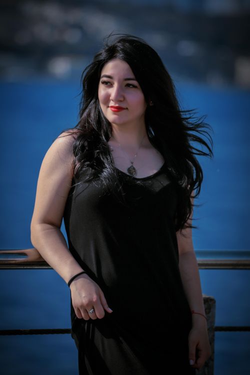 Free Attractive brunette in stylish black dress standing near railing against blurred sea and looking away dreamily Stock Photo