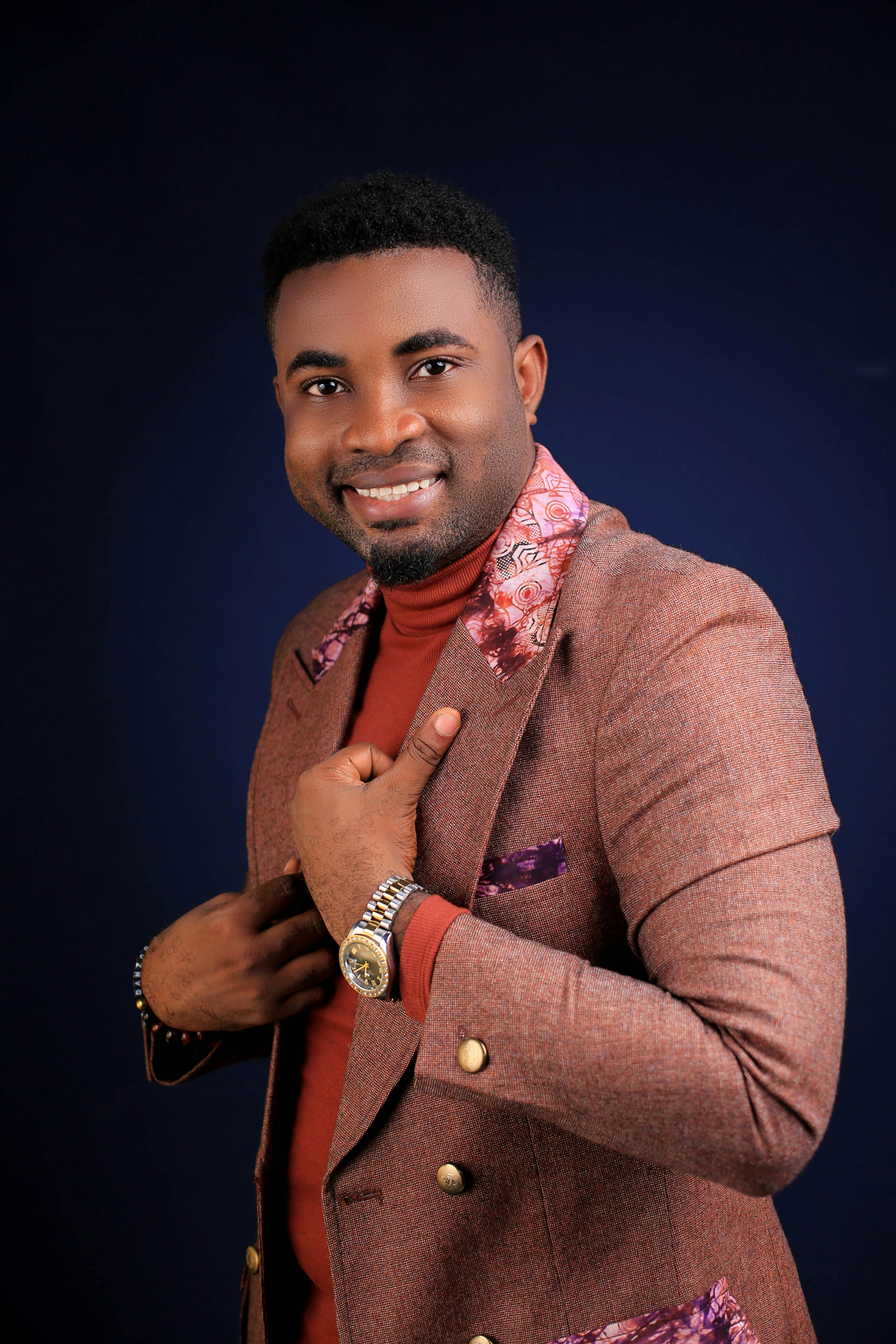 Happy African American plump male with black curly hair adjusting beige formal jacket and looking at camera with bright friendly toothy smile on blue background