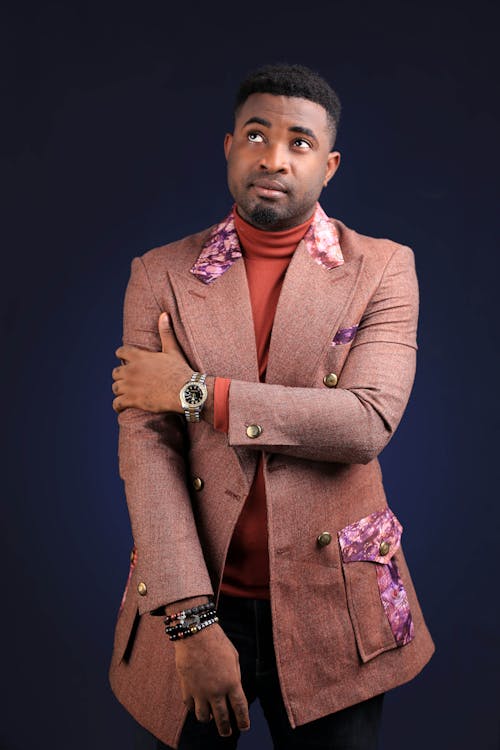 Free Pensive African American businessman in beige formal jacket and wristwatch and bracelet on hands looking up on blue background Stock Photo