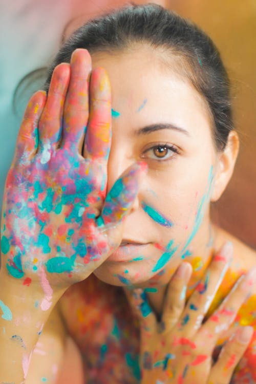 Free Unemotional young shirtless female artist with golden eyes and dark hair tied up covering eye with painted arm and looking at camera on multi colored blurred background Stock Photo