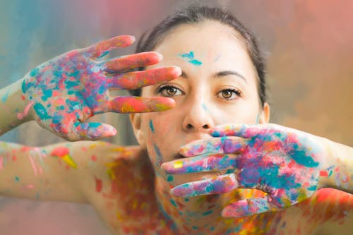 Serious slim young topless female artist with golden eyes and palms painted with multicolored dye looking at camera on blurred background