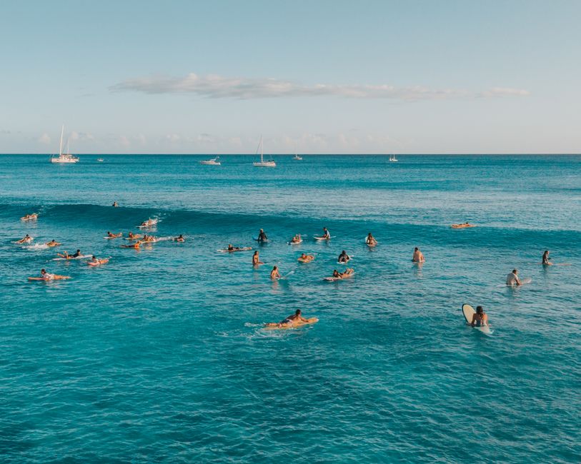 Aerial view of surfers enjoying the waves in the beautiful ocean of Hawaii, with sailboats in the distance.