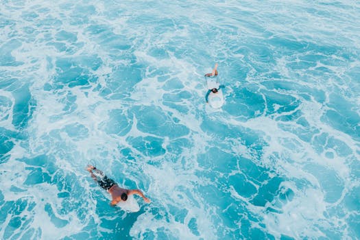 Two surfers paddle through vivid blue ocean waves, captured from above in Hawaii.