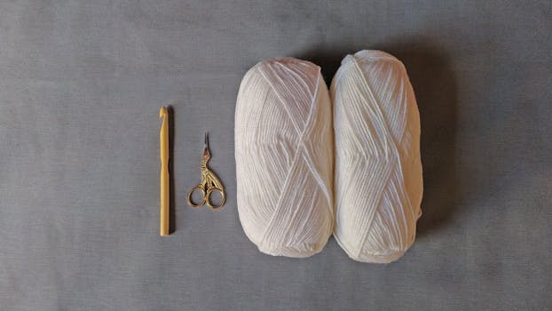 A minimalist flat lay featuring a crochet hook, scissors, and white yarn on a gray background.
