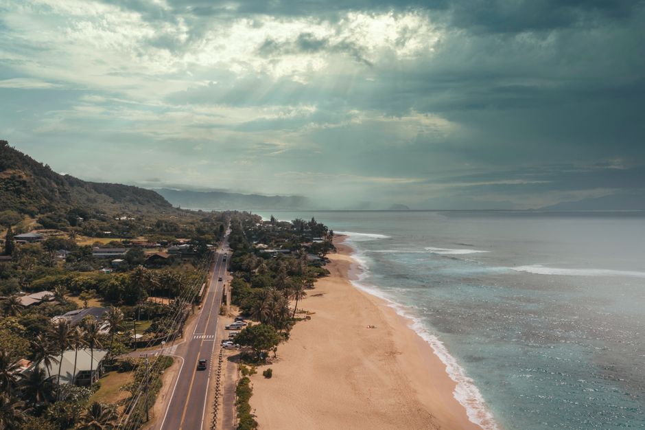Photo by Jess Loiterton Stunning aerial photograph of Oahu's North Shore, capturing the beach, road, and lush landscape under dramatic skies.