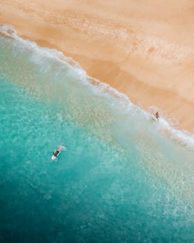 Drone captures turquoise waters meeting sandy shores, two surfers enjoying the ocean.