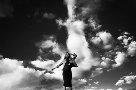 A fashionable woman poses confidently against a dramatic backdrop of clouds. Black and white photography enhances the scene.